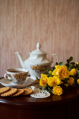 Still life and yellow roses, teapot with cup and round cookies on brown table