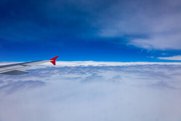 The wing of the plane flies over white clouds, the background is blue sky, view through the window.