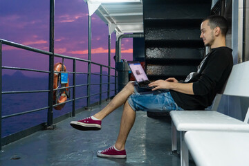 A young man works on a laptop on the deck of a ship. Remote work. Always in touch.