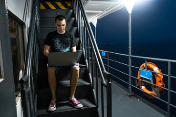 A young man works on a laptop on the deck of a ship. Remote work. Always in touch.