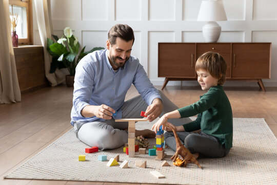 Happy father playing with little son, sitting on warm floor with underfloor heating, smiling dad and cute boy having fun, building tower from colorful wooden blocks, enjoying leisure time together