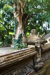 Beautiful, ancient stone fence with carved columns near the temple on the island of Bali. There is a large green tree in the background.