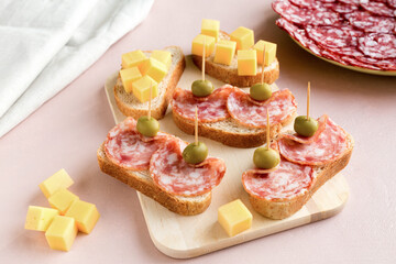 Sandwiches of smoked sausage and white bread on a pink paper background. The dish is decorated with green olives. There is a plate of sliced sausage on the back.