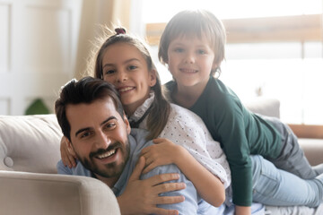Fototapeta na wymiar Head shot portrait happy father relaxing on cozy couch with adorable children, cute little son and daughter lying on dad back, looking at camera, family enjoying lazy weekend, leisure time at home