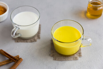 In one glass cup, ordinary milk, in the other, Golden milk an Indian hot drink with turmeric root powder. Next to it are the ingredients curcuma, honey and cinnamon sticks. Gray background.