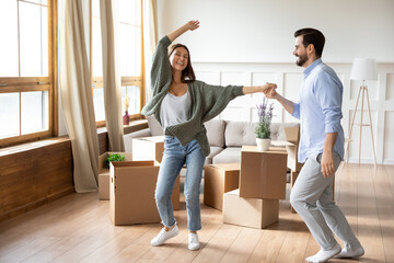 Fototapeta na wymiar Happy young wife and husband dancing in modern living room with cardboard boxes with belongings, excited family celebrating moving day, satisfied customers relocating into new apartment, mortgage