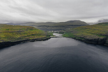 Small bay in a fjord aerial panorama. Overcast sky, deep blue ocean and green hills. Faroe Islands, Denmark.