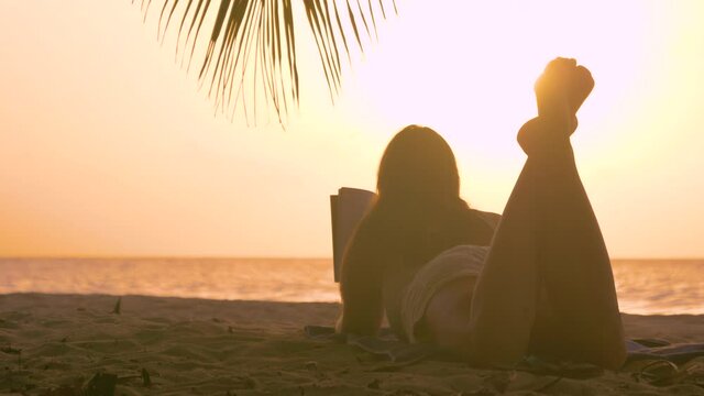 CLOSE UP, LENS FLARE: Unrecognizable tourist girl enjoys a sunny summer evening on the scenic beach by reading a good book. Young woman on vacation in Barbados reads on a sandy beach at sunset.