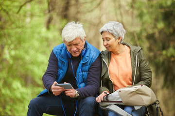 Portrait of active senior couple using smartphone while resting during hike in autumn forest, copy space