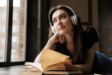 Photo of young woman wearing headphones lying on wooden floor with book