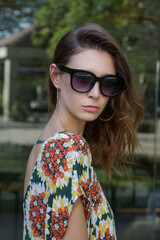 Outdoor portrait of stylish fashionable woman in colorful dress and big black sunglasses. Casual summertime trendy outfit. Headshot of young brunette on the street looking at camera.

