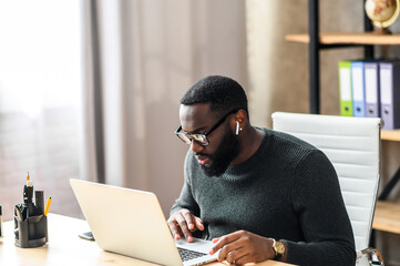 Concentrated black guy in smart casual wear using laptop for work or studying. African-American guy...