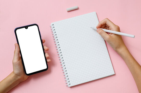 The right hand holds a pen over a notebook sheet. Left hand holds a mobile phone with a blank display