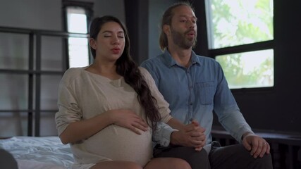 4K Beautiful pregnant woman and her husband sitting on the bed and do practicing breathing exercise together for preparing baby childbirth. Family relationship and maternity healthcare concept.