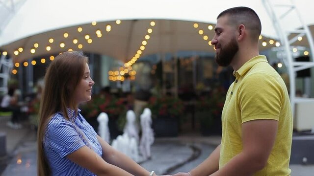 Loving guy and girl holding hands opposite each other smiling hugging on street, defocused background of fountain and restaurant in evening. Romantic date of young couple. Concept of love relationship