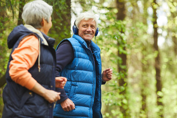Side view portrait of active senior couple running in forest lit by sunlight and looking at each...