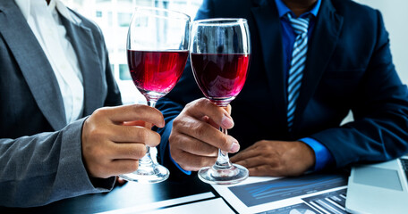 Two business executives people, businesswomen, and businessman in a suit is bumping into a glass of wine celebrating success at the office desk. After the meeting
