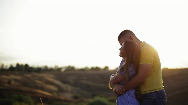 Tall young bearded guy in polo shirt hugs behind pretty girl in striped dress and hold hands in field against defocused background of sunset in evening. Romantic date in nature outside couple in love