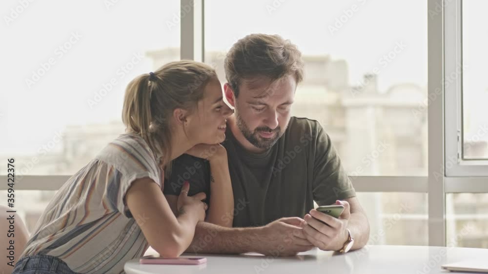 Wall mural A lovely young couple man and woman are spending time together watching something on the smartphone while sitting at the table at home