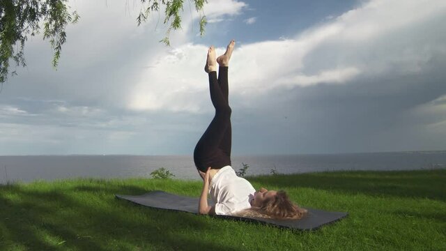 Young fit woman practice yoga on coast near the lake or sea. Woman doing Supported Shoulder Stand Pose