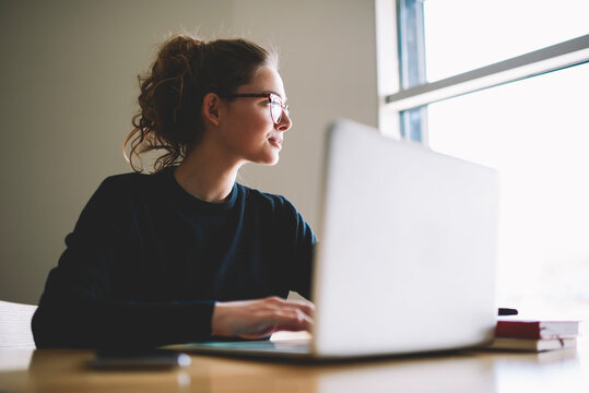 Charming Business Woman In Stylish Optical Spectacles Sitting Indoors At Table And Working Freelance At Laptop Computer With High Speed 4G Internet Connection While Looking Out Of Window