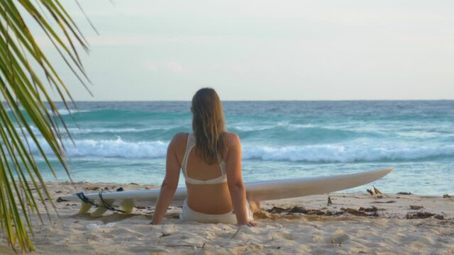 SLOW MOTION, CLOSE UP, DOF: Unrecognizable young female traveler on a surfing trip in the Caribbean enjoys a calm summer morning on a beautiful sandy beach. Surfer girl watches the waves at sunrise.