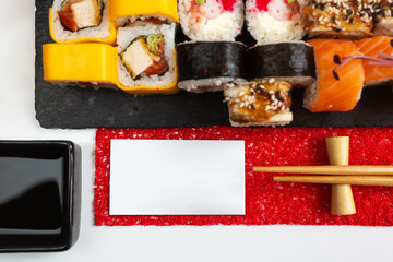 Different types of sushi rolls on a dark stone board. Wooden chopsticks with card. Blank white card for text.