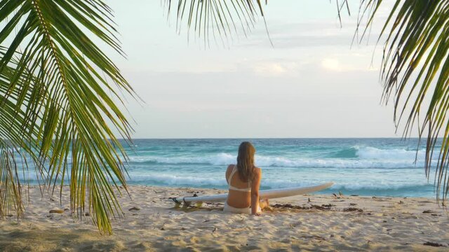 SLOW MOTION: Unrecognizable young woman sits on the sandy beach with a surfboard and watches the ocean at sunset. Female traveler enjoys a relaxing moment on the tropical white sand shore of Barbados.