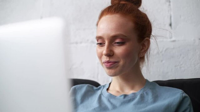 Close-up of face of redhead young businesswoman with emotion of joy and success working on laptop looking on screen of monitor. Cheerful business lady is chatting on computer at remote workplace.
