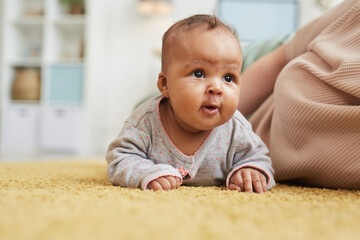 Warm-toned portrait of cute mixed-race baby looking up while crawling on fluffy yellow carpet at home, copy space