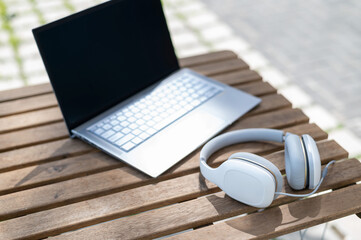 Close-up of a laptop and white big headphones on a wooden table on a summer terrace. No people. Cafe at the outdoors. Freelancer workspace.