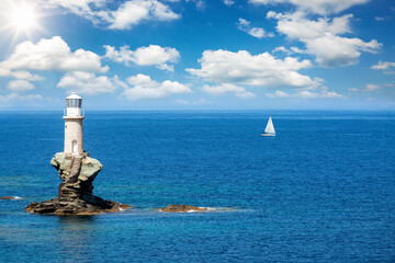 A lighthouse on a steep rock in calm, blue sea with a passing by sailing boat at the horizon,...