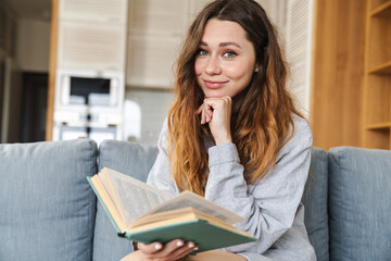 Photo of cheerful woman smiling and reading book while sitting on couch