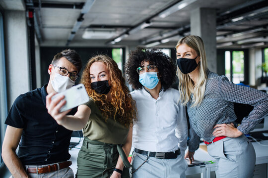 Young people with face masks back at work in office after lockdown, taking selfie.
