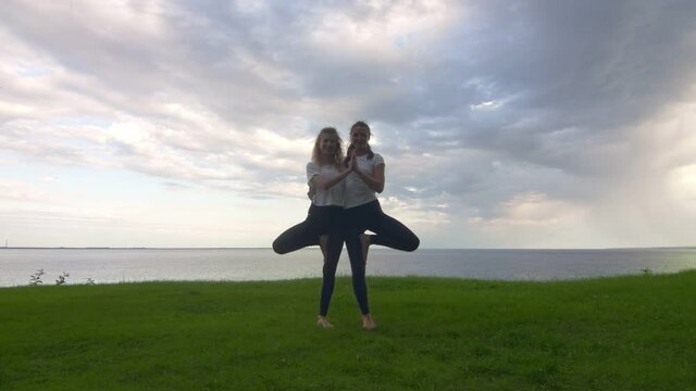 Two women in activewear pposing on the camera and showing together yoga Tree pose Vrksasana Vriksasana