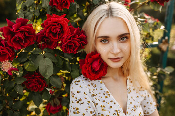 Young woman portrait with freckles and long blonde hair, standing next to a bush with burgundy roses in sun light, outdoors, looking to camera.