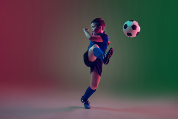 Fototapeta na wymiar Attacking. Teen male football or soccer player on gradient background in neon light. Caucasian boy training, practicing on the run, in jump. Concept of sport, competition, winning, motion, action.