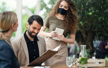 Waitress with face mask serving happy couple outdoors on terrace restaurant.