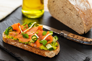 Open faced sandwich with smoked salmon, avocado, apple, zucchini, pumpkin, basil and chives on toasted sourdough bread. With knife, bread loaf and olive oil on slates.