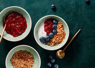 Top view of breakfast with granola, yoghurt, strawberry jam, chia seeds and blueberry on a dark green table.