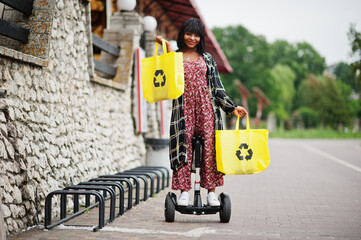Beautiful african american woman using segway or hoverboard. Black girl with yellow  cloth eco bags recycling symbol.