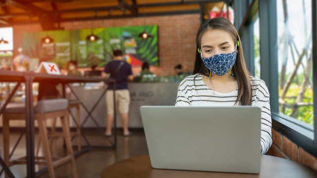 New Normal And Social Distancing After Covid Epidemic Concept.young Smart Asian Female Watching Laptop Meeting Business Team Or Webinar Course Online With Face Shield Or Mask Protection At Coffee Shop