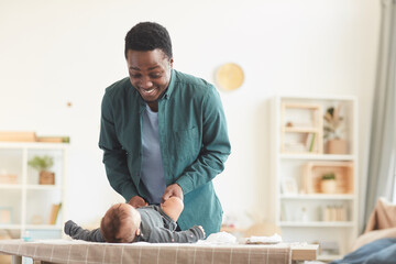 Warm-toned portrait of loving African-American father playing with cute baby boy while dressing him...