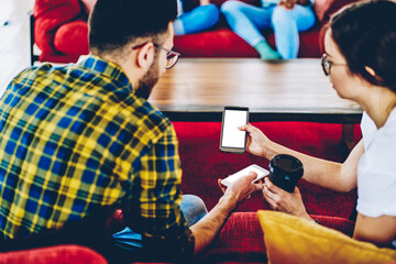 Millennial hipster girl showing smartphone with blank screen for your internet content to friend sitting in coworking space.Male and female bloggers watching video on website on cellular using 4G