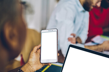 Selective focus on modern smartphone device with copy space display in woman's hands.Cropped view of hipster girl holding telephone with blank screen and sharing media files on laptop computer