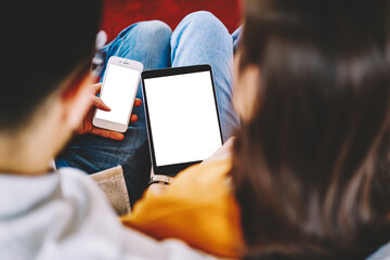 Cropped back view of young marriage using modern smartphone and touch pad with blank screen area...