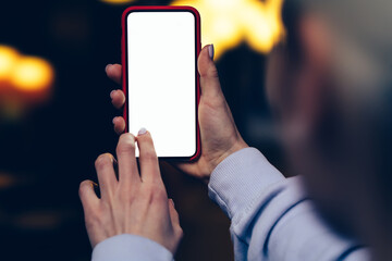 Rear view of female holding in hands mobile phone touching clear display with finger against gold shinning bokeh lights. Woman keeps modern smartphone with blank mockup copy space screen for advertise