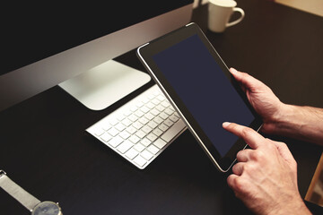 Cropped image of man's hands using portable digital tablet with blank copy space screen for your text message or promotional content against freelancer workplace with pc, wireless keyboard, cup of tea