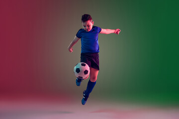 Overcoming. Teen male football or soccer player on gradient background in neon light. Caucasian boy training, practicing on the run, in jump. Concept of sport, competition, winning, motion, action.