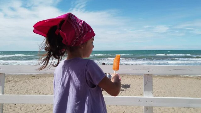 baby girl child eat popsicle at the beach bar on wind sea with pink bandana